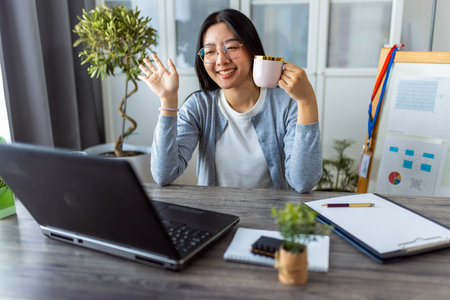 A young Thai woman enjoys a cup of coffee while participating in an online business video call from her home office.の写真素材