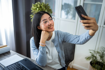Thai woman is seated at her desk in a cozy home office, working on her computer while pausing to take a selfie with her smartphone.の写真素材