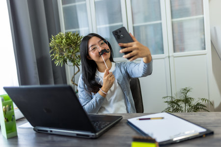 A young Thai woman sits at her desk in a home office environment, playfully wearing fake mustaches as she takes a selfie with her smartphone.の写真素材