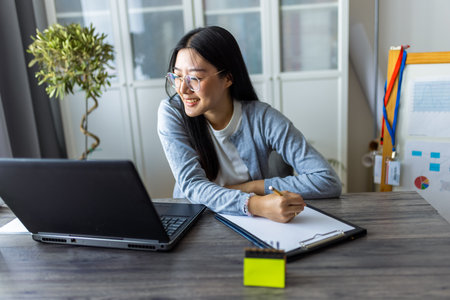 Thai woman sits at a desk in a comfortable home environment, focused on her computer as she works remotely.の写真素材