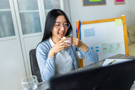 A young Thai woman enjoys a cup of coffee while participating in an online business video call from her home office.の写真素材