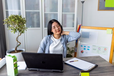 A young Thai woman working from home on her computer looks exhausted and stretches her arms and head to relieve tension.の写真素材