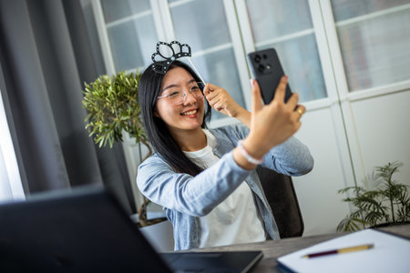 A young Thai woman is having fun while taking a selfie wearing a playful fake crown.の写真素材