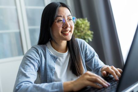 Thai woman sits at a desk in a comfortable home environment, focused on her computer as she works remotely.の写真素材