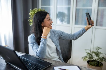 Thai woman is seated at her desk in a cozy home office, working on her computer while pausing to take a selfie with her smartphone.の写真素材
