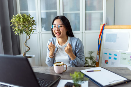 A young Thai woman enjoys a cup of coffee while participating in an online business video call from her home office.の写真素材