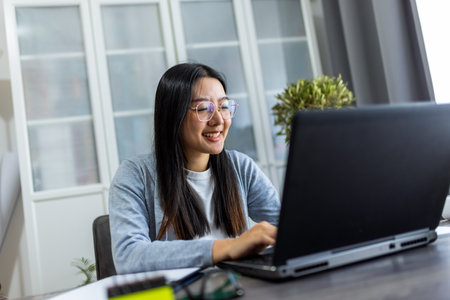 Thai woman sits at a desk in a comfortable home environment, focused on her computer as she works remotely.の写真素材
