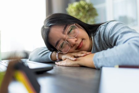 A young Thai woman working from home on her computer looks extremely tired and rests her head on the table, showing signs of fatigue and exhaustion.の写真素材