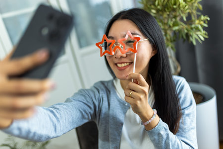 A young Thai woman sits at her desk wearing playful star-shaped glasses while taking a selfie with her smartphone.の写真素材