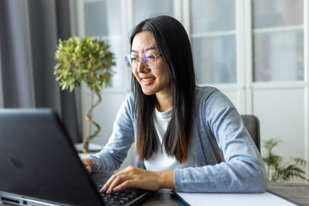 Thai woman sits at a desk in a comfortable home environment, focused on her computer as she works remotely.の写真素材