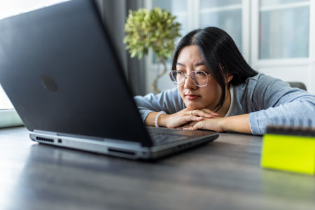 A young Thai woman working from home on her computer looks extremely tired and rests her head on the table, showing signs of fatigue and exhaustion.の写真素材