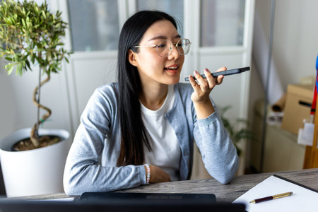 Thai woman sits at a desk in a modern home setting, multitasking as she works on her computer and sends a voice message on her smartphone.の写真素材