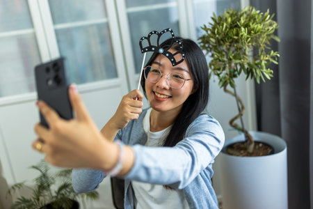 A young Thai woman is having fun while taking a selfie wearing a playful fake crown.の写真素材