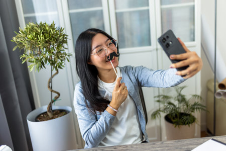 A young Thai woman sits at her desk in a home office environment, playfully wearing fake mustaches as she takes a selfie with her smartphone.の写真素材