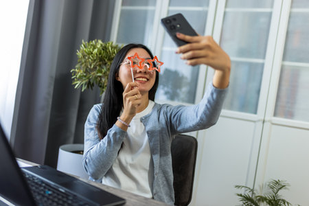 A young Thai woman sits at her desk wearing playful star-shaped glasses while taking a selfie with her smartphone.の写真素材