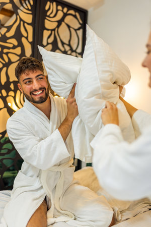 A joyful couple dressed in soft white bathrobes has a playful pillow fight in bed, laughing and enjoying each otherâs company.の写真素材