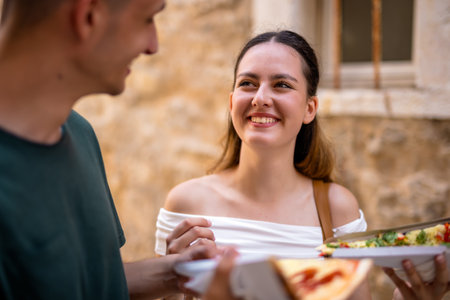 A happy young couple enjoys their summer holiday exploring a charming old town. They walk together casually while sharing slices of pizza.の写真素材
