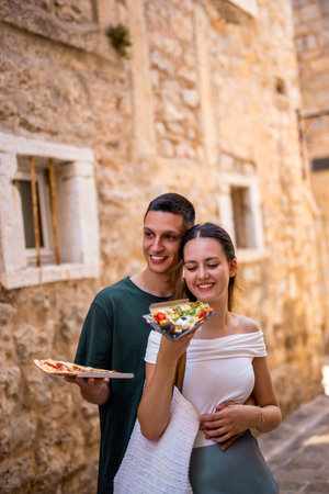 A young couple enjoys a romantic summer holiday in a charming old town. The boyfriend embraces his girlfriend from behind as she happily eats a slice of pizza.の写真素材
