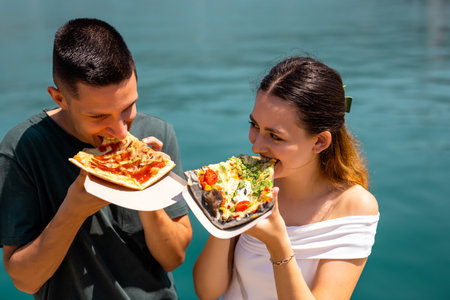A young couple enjoys a relaxed summer holiday moment on the shore by the sea. They share slices of pizza while talking and smiling, dressed in casual summer clothes.の写真素材