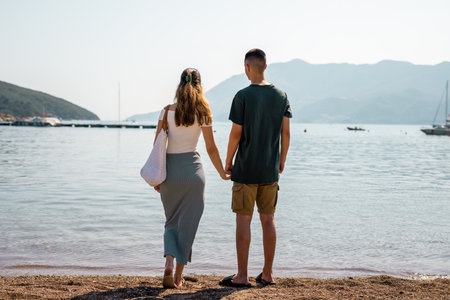 Young couple is holding hands with the sea behind them on the beach. The view is from the back.