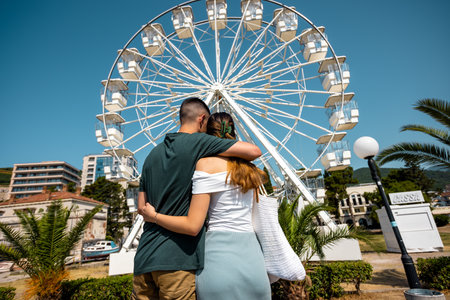 A young couple stands by the shore on a sunny summer day, hugging each other and gazing at a Ferris wheel in the distance.の写真素材