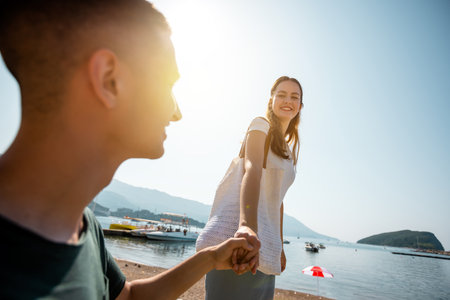 A young couple enjoying a summer day by the shore. The girlfriend carefully walks along a low ledge or uneven surface, while her boyfriend gently holds her hand to support her and keep her from falling.の写真素材