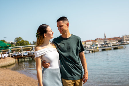 A young couple stands close by the sea on a sunny day, with the boyfriend gently holding his...