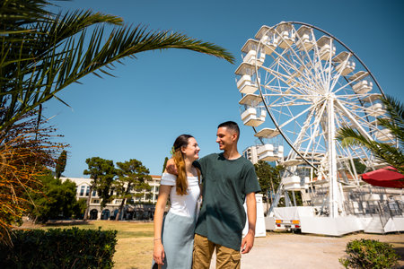 Young couple enjoying a romantic summer day by the shore, standing in front of a Ferris wheel at a seaside amusement park. They are dressed in light, summer clothes, holding each other affectionately and engaged in conversation.の写真素材