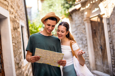 A young couple wanders through a charming seaside old town, holding a map as they explore the historic streets. The boyfriend wears a hat, and both are dressed in light summer clothes.の写真素材