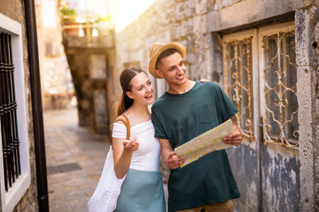 A young couple wanders through a charming seaside old town, holding a map as they explore the historic streets. The boyfriend wears a hat, and both are dressed in light summer clothes.の写真素材