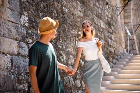 A young couple enjoys their summer holiday exploring the historic charm of an old city. The girlfriend carefully walks down the stone stairs along the ancient city walls, while her boyfriend gently holds her hand to support and guide her.の写真素材