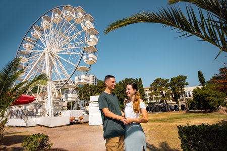Young couple enjoying a romantic summer day by the shore, standing in front of a Ferris wheel at a seaside amusement park. They are dressed in light, summer clothes, holding each other affectionately and engaged in conversation.の写真素材