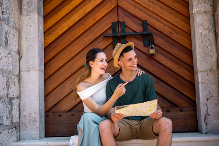 A young couple sits together by the ancient gate of a historic old city during their summer holiday, examining a map as they plan their next adventure.の写真素材