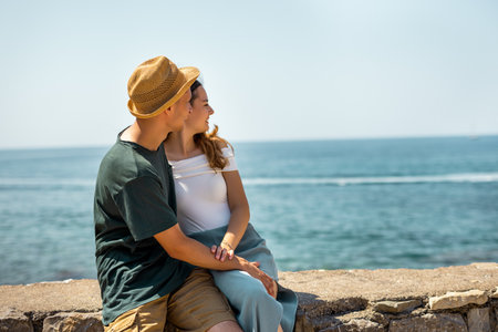 A romantic couple enjoys a peaceful summer holiday moment, sitting on the ancient stone walls of a historic old city. They hold each other closely and gaze at the sea.の写真素材