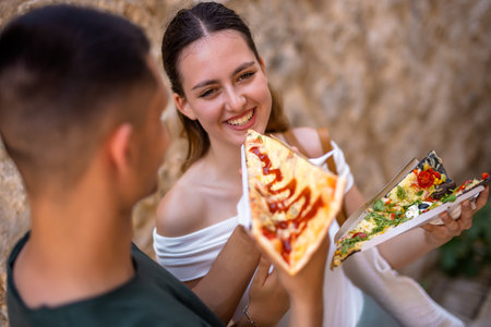 A joyful young couple enjoys their summer holiday in a charming old town. The boyfriend lovingly feeds his girlfriend a slice of pizza as they share a fun, romantic moment together.の写真素材