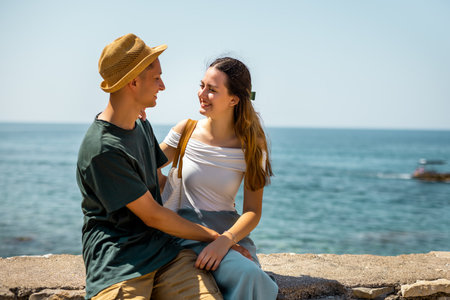 A young couple shares a romantic moment during their summer holiday, sitting on the ancient stone walls of a historic old city. They hold each other affectionately and gaze into each otherâs eyes, with the sea stretching out in the background.の写真素材