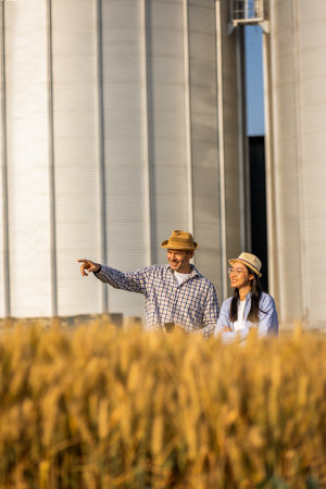 Two workers stand in a wheat-covered area in front of large silos, they look at something in the distanceの写真素材