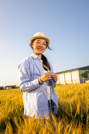 A Thai female farmer stands in a golden wheat field holding wheat stalks and smiling
