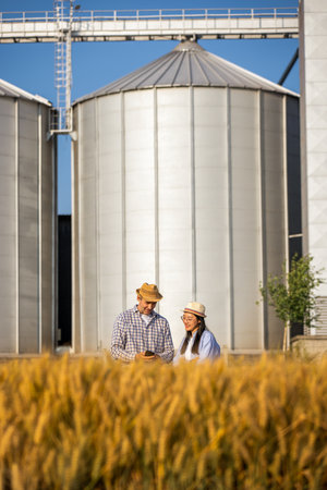 A Thai woman and an older male worker stand in a pile of wheat in front of large silos, using a smartphone together.の写真素材
