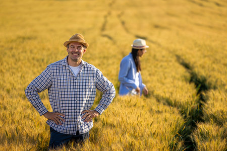 A senior man smiles confidently at the camera while standing in a golden wheat field, with a Thai female farmer working diligently in the background.の写真素材
