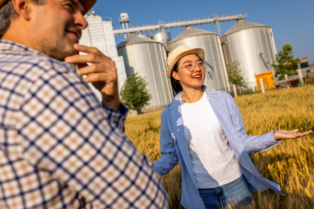 Two workers stand in a wheat-covered area in front of large silos, actively discussing upcoming tasks.の写真素材