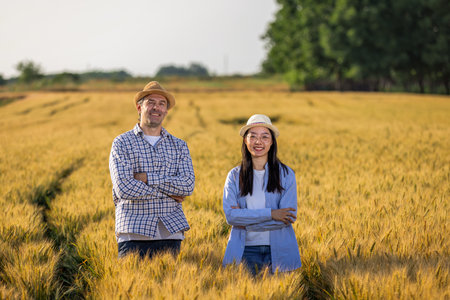 A Thai woman and an older male farmer stand confidently with crossed arms and smiling faces in a golden wheat field.の写真素材