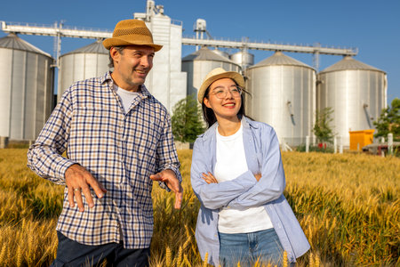 Two workers stand in a wheat-covered area in front of large silos, actively discussing upcoming tasks.の写真素材