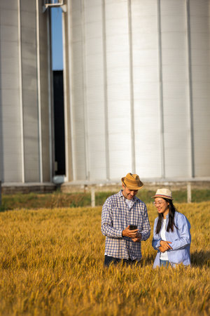 A Thai woman and an older male worker stand in a pile of wheat in front of large silos, using a smartphone together.の写真素材