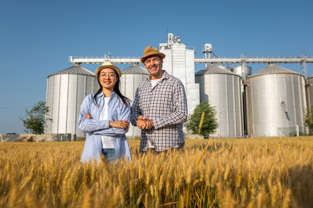 Two workers stand in a wheat-covered area in front of large silos, actively discussing upcoming tasks.の写真素材