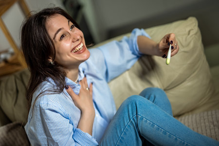 A young woman is sitting in her living room, smiling with joy and excitement as she looks at a positive pregnancy test.の写真素材