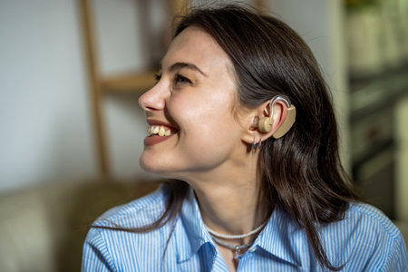 A cheerful young woman is sitting in her living room, smiling and enjoying the moment, proudly wearing a hearing device on her ear.の写真素材