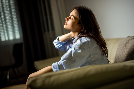 A young woman is sitting in her living room looking sad and emotionally overwhelmed, portraying a moment of depression.の写真素材