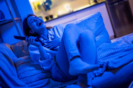 A young woman sits comfortably in her living room bathed in blue light from the TV, laughing joyfully as she watches a comedy movie, enjoying a fun and relaxed moment at home.の写真素材
