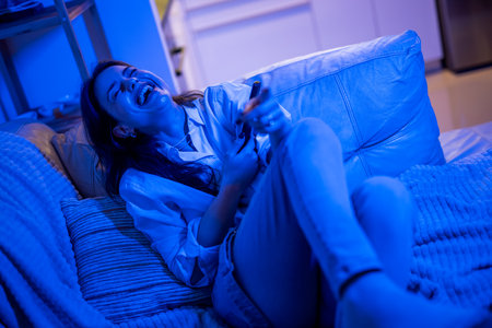 A young woman sits comfortably in her living room bathed in blue light from the TV, laughing joyfully as she watches a comedy movie, enjoying a fun and relaxed moment at home.の写真素材